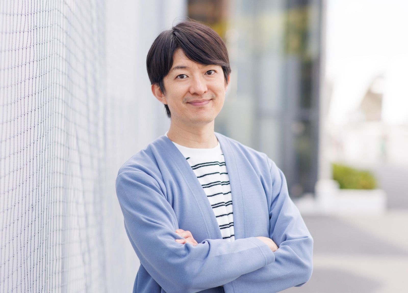 白いフェンスの前で腕を組み、青いカーディガン姿で微笑む澤田智洋